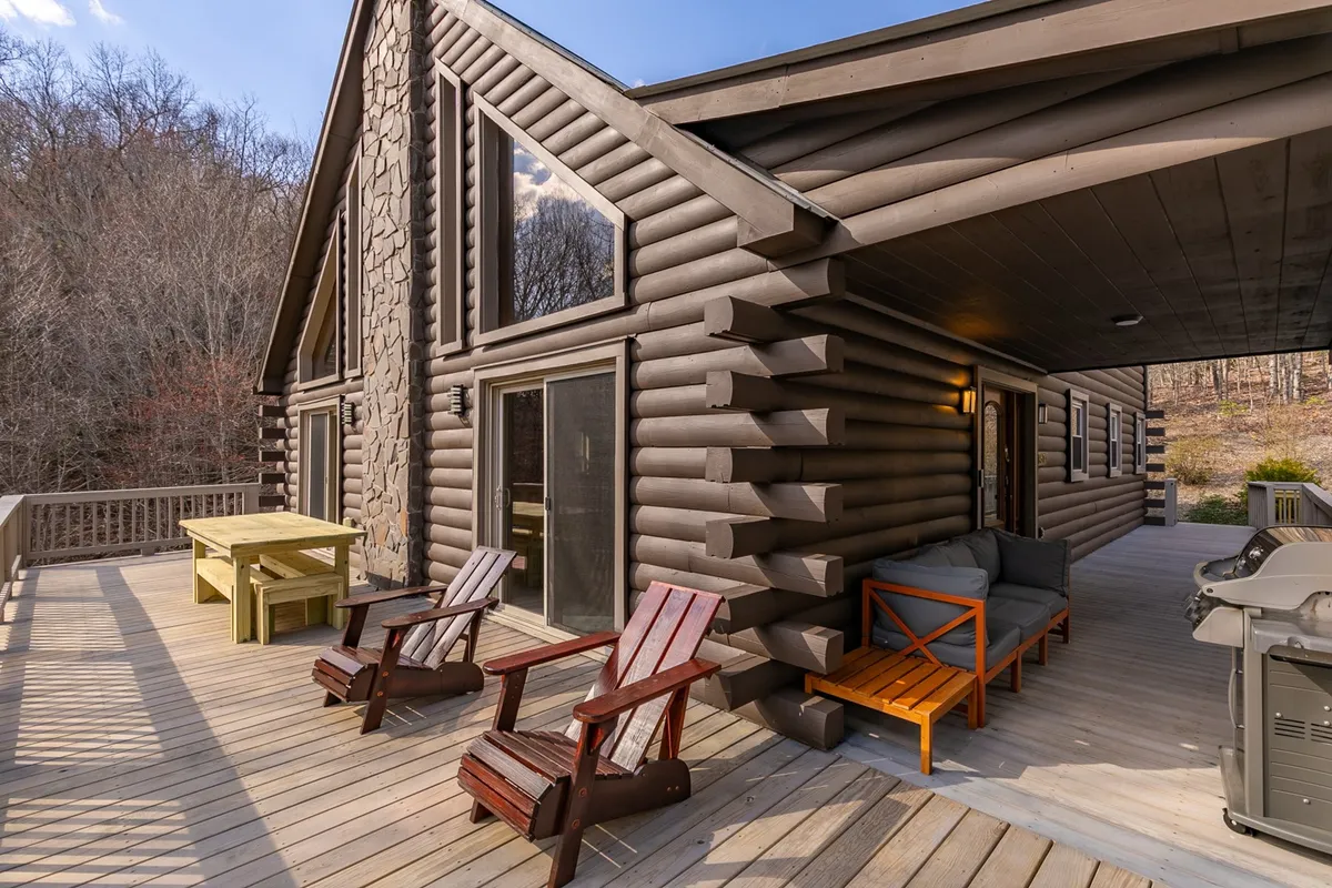 A welcoming exterior shot of the log cabin with large windows and a spacious deck outfitted with outdoor seating. The deck faces lush surroundings, suggesting an inviting spot for gatherings, al fresco dining, and relaxation with pets.