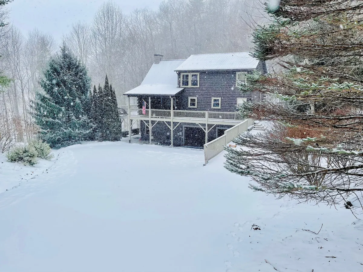 A tranquil winter exterior showing the house, expansive deck, and snow-covered yard surrounded by trees. The deck features seating areas ideal for relaxing outdoors in every season, and the snowy landscape hints at opportunities for pets and guests to enjoy the natural setting.
