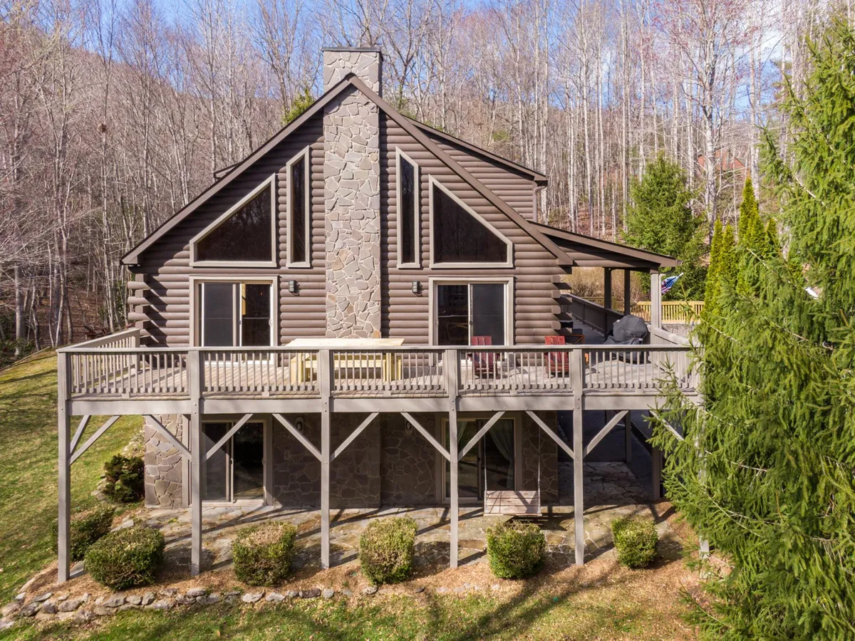 The hero image showcases a charming log cabin-style home with a robust stone chimney, a spacious deck furnished for relaxation and outdoor dining, and beautifully landscaped gardens. The scene radiates warmth and coziness, hinting at sunrise views from the deck and inviting guests to imagine gathering with loved ones or enjoying peaceful nature moments.