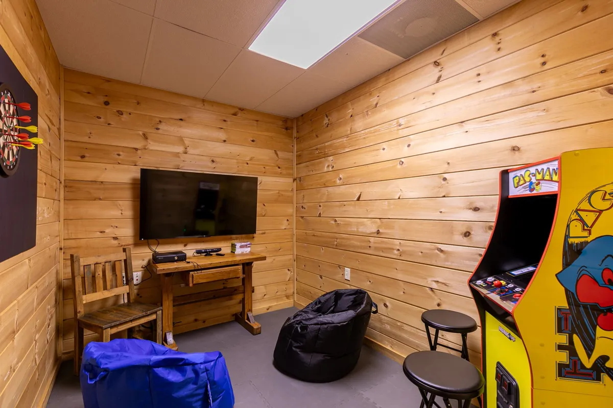 The focal point of this scene is a Pac-Man arcade machine, flanked by a dartboard, bean bag chairs, and a TV. The relaxed arrangement encourages group play and laid-back lounging, ideal for family gaming sessions and movie marathons. The presence of multiple gaming options—arcade, darts, and video—echoes the section's emphasis on versatility and fun.