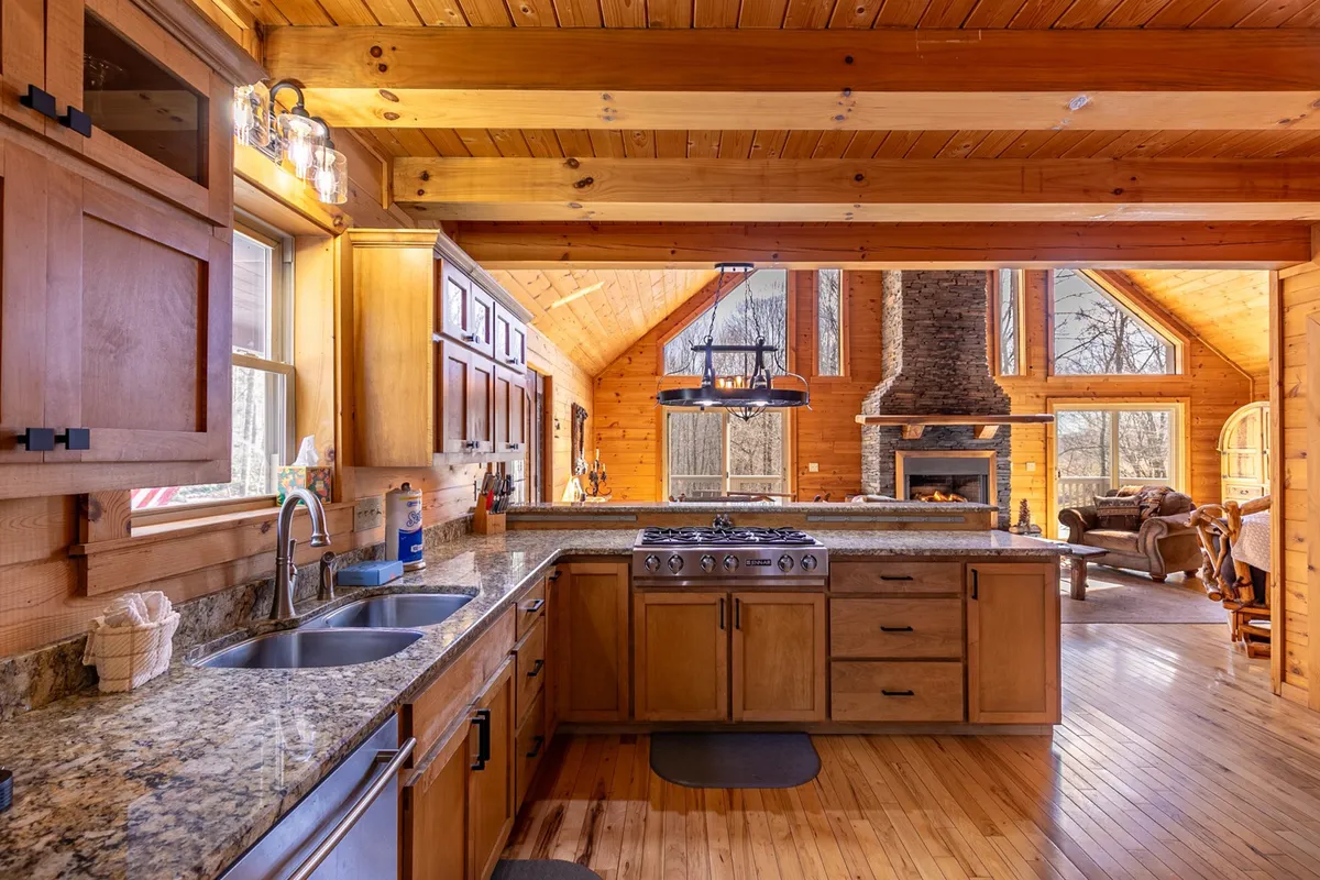 A view merging the kitchen and living area, with granite countertops, stainless steel appliances, a stone fireplace, and large windows. The kitchen opens directly to the main living space, highlighting the modern, fully stocked kitchen and the open-concept design.