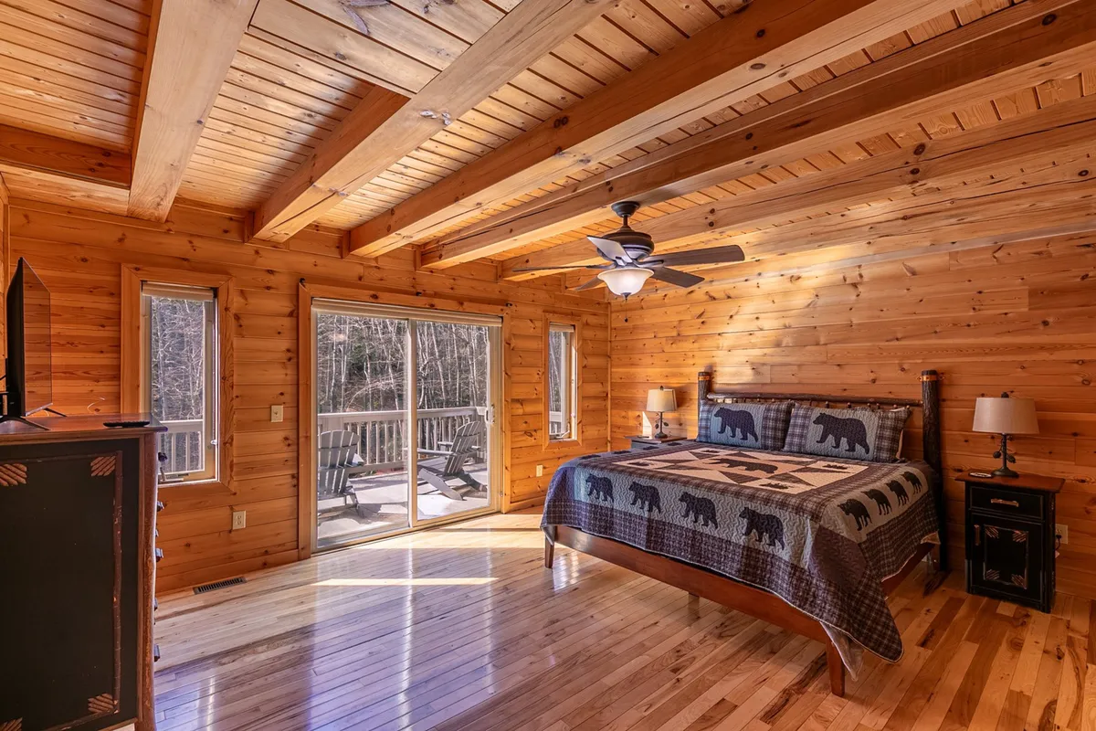 A warm and rustic bedroom featuring a bed with bear-themed bedding, large windows offering a view of the outdoors, and wooden beams and walls. The space feels inviting and tranquil, ideal for relaxation or enjoying a morning coffee on the deck.