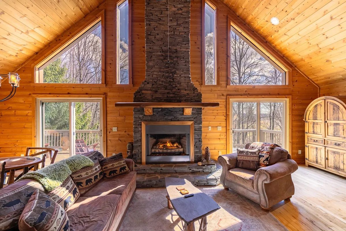 A cozy interior space with a prominent stone fireplace, large windows revealing the outdoors, rustic wooden beams, and inviting furniture. The room is filled with warm light, inviting relaxation after a day of outdoor activities.