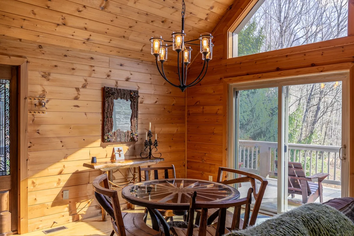 A warm dining area with a wooden table, a chandelier, large windows letting in natural light, and wood-paneled walls. This inviting space is set up for intimate meals, family board games, or special celebrations, embodying the communal spirit of the home.