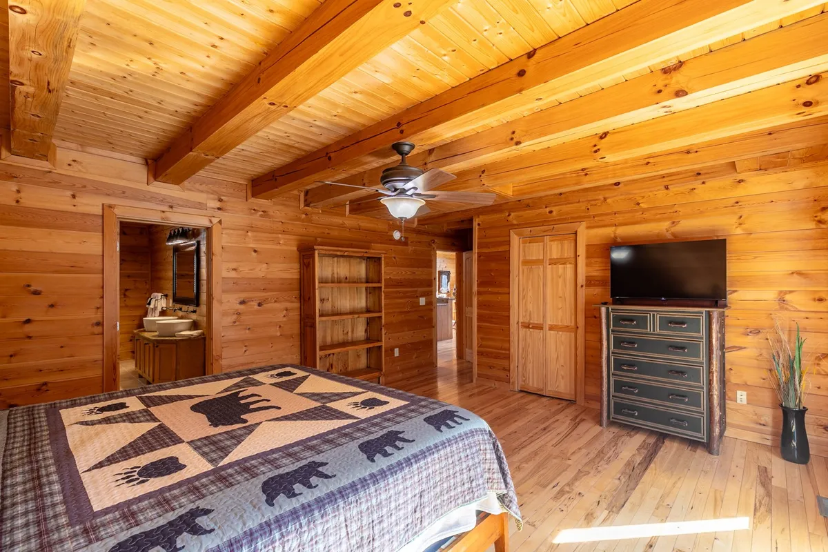 A bedroom with a patterned quilt on the bed, exposed wooden ceiling beams, a dresser with a TV, and an open doorway leading to a bathroom. Natural light creates a welcoming and restful atmosphere, perfect for unwinding after a day of adventure.