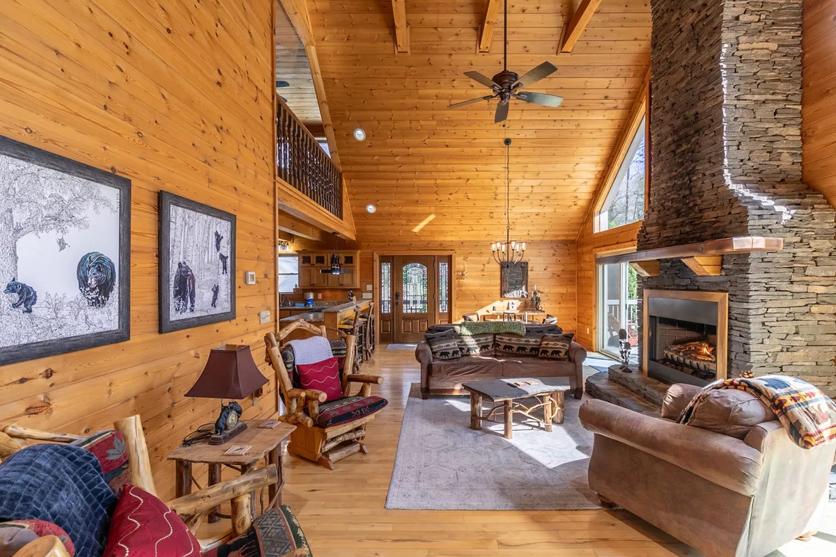 A spacious living room with a stone fireplace, large windows, wooden beams, and comfortable seating. The expansive windows frame the surrounding landscape, filling the space with light and offering direct views of nature.