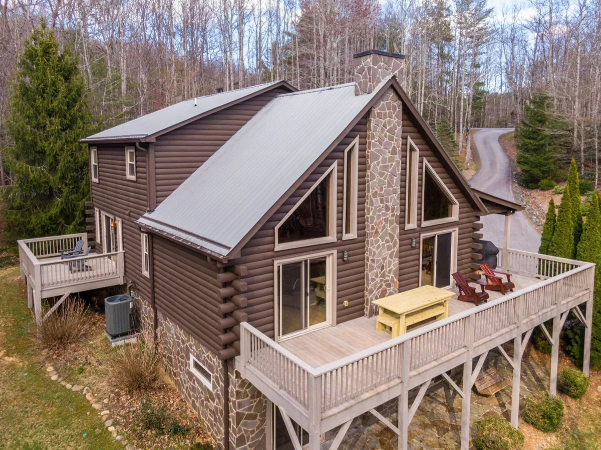 An exterior view highlighting the wooden deck with rocking chairs, a stone chimney, and large windows, framed by surrounding trees. The scene exudes tranquility and showcases the deck as a central hub for family gatherings and quiet enjoyment of the mountain air.
