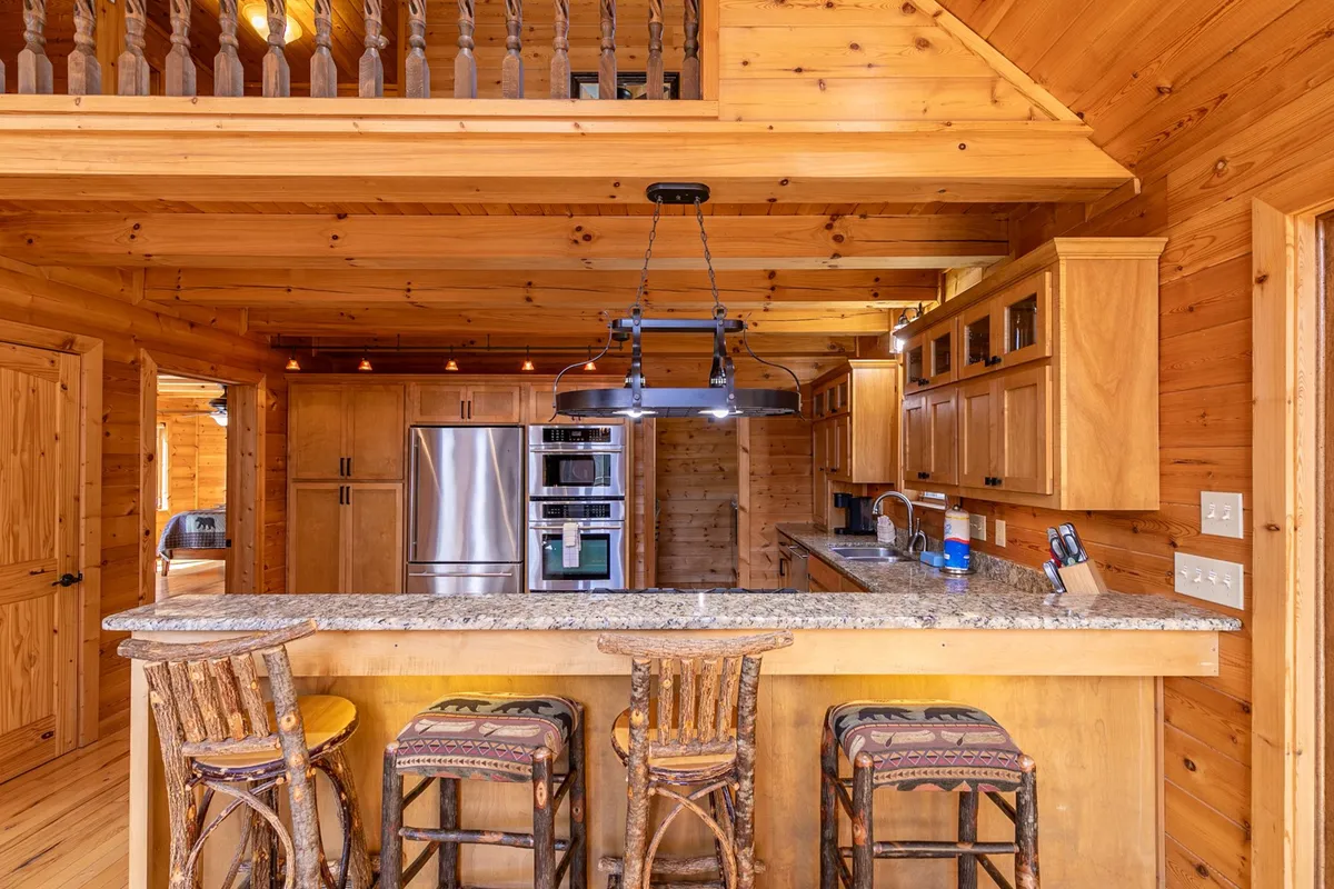 A close-up view of the kitchen’s granite countertop with wooden bar stools, open shelving, and visible kitchen appliances. The layout encourages socializing and showcases the breakfast bar, ideal for casual conversations or coffee breaks.