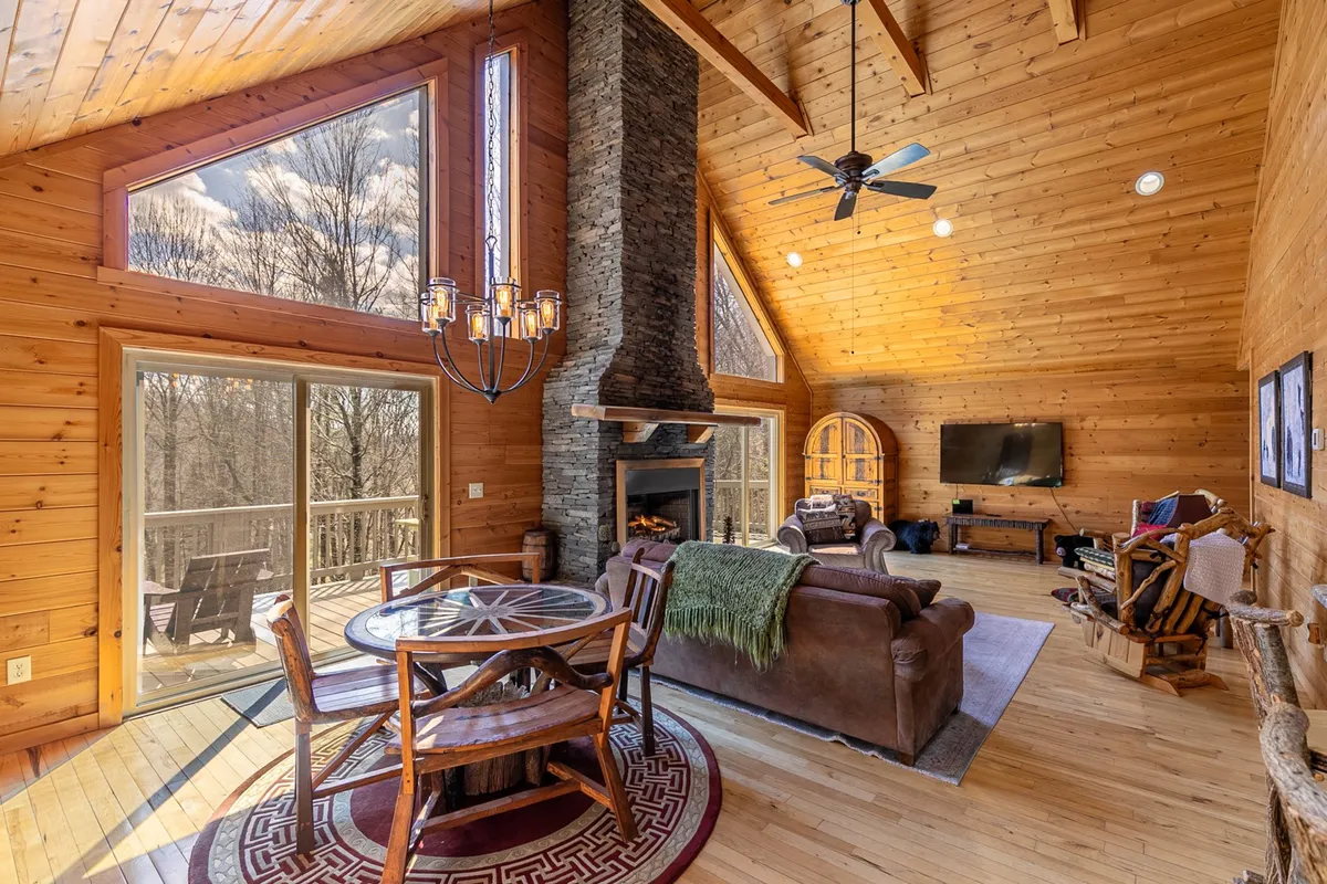 A spacious great room featuring a stacked stone fireplace, large windows that flood the space with natural light, a leather sofa, a wooden dining table, and exposed wooden beams. The open-plan layout shows the seamless connection between the living and dining areas, creating an inviting atmosphere for family gatherings and celebrations.