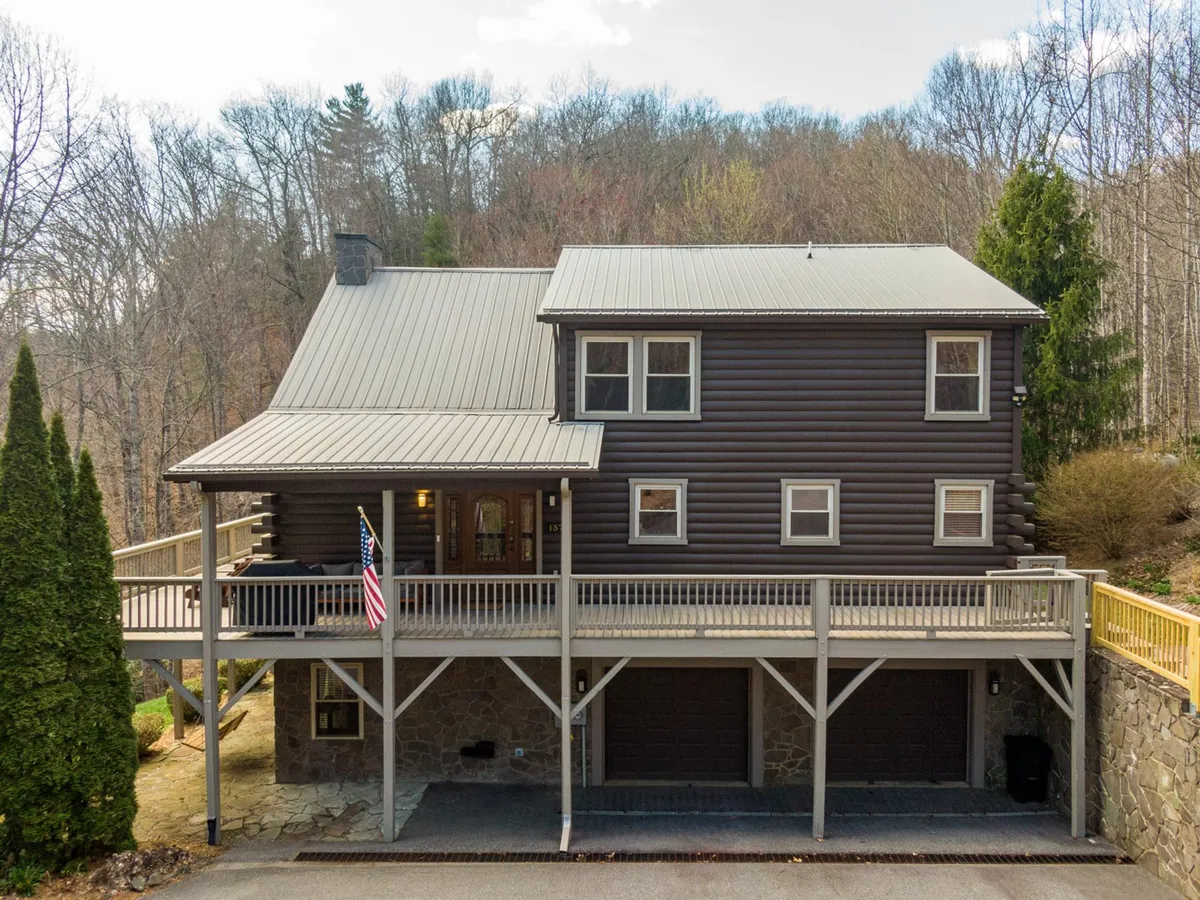 A log cabin structure framed by trees, with a spacious front porch and a waving American flag, offers a picturesque and welcoming exterior view. The natural setting and spacious porch suggest a perfect base for group adventures and relaxing together after exploring nearby attractions.